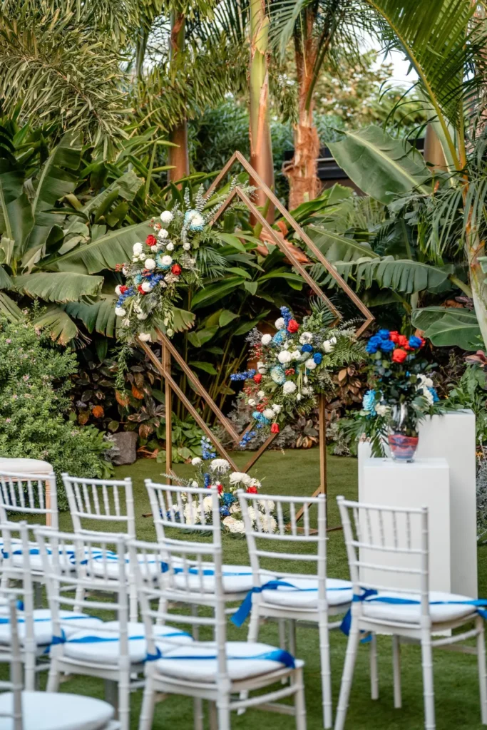 Vista desde los asientos hacia el altar con arco floral, pedestal de flores y sillas con lazos azules