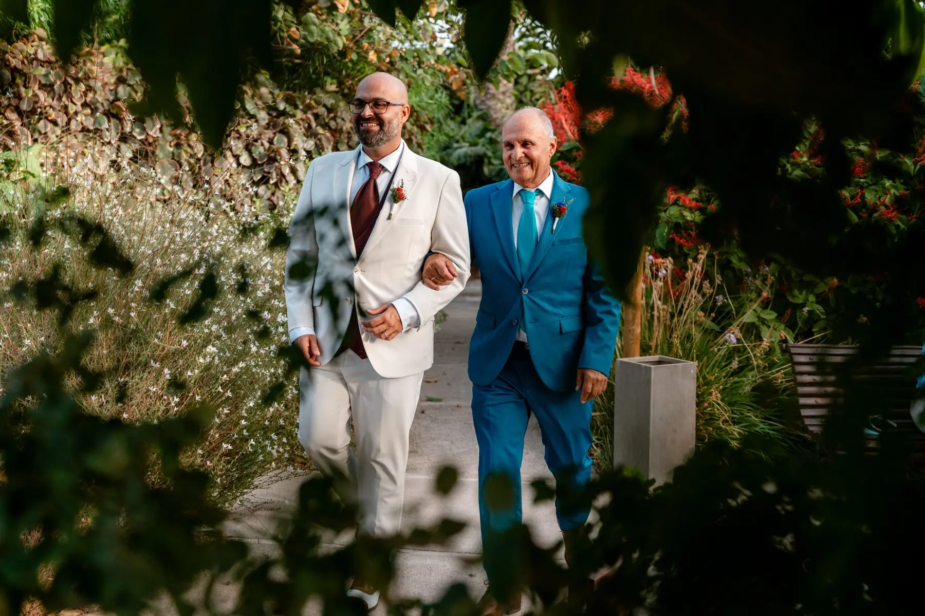 Novio con traje blanco y corbata burdeos caminando del brazo de su padre entre vegetación